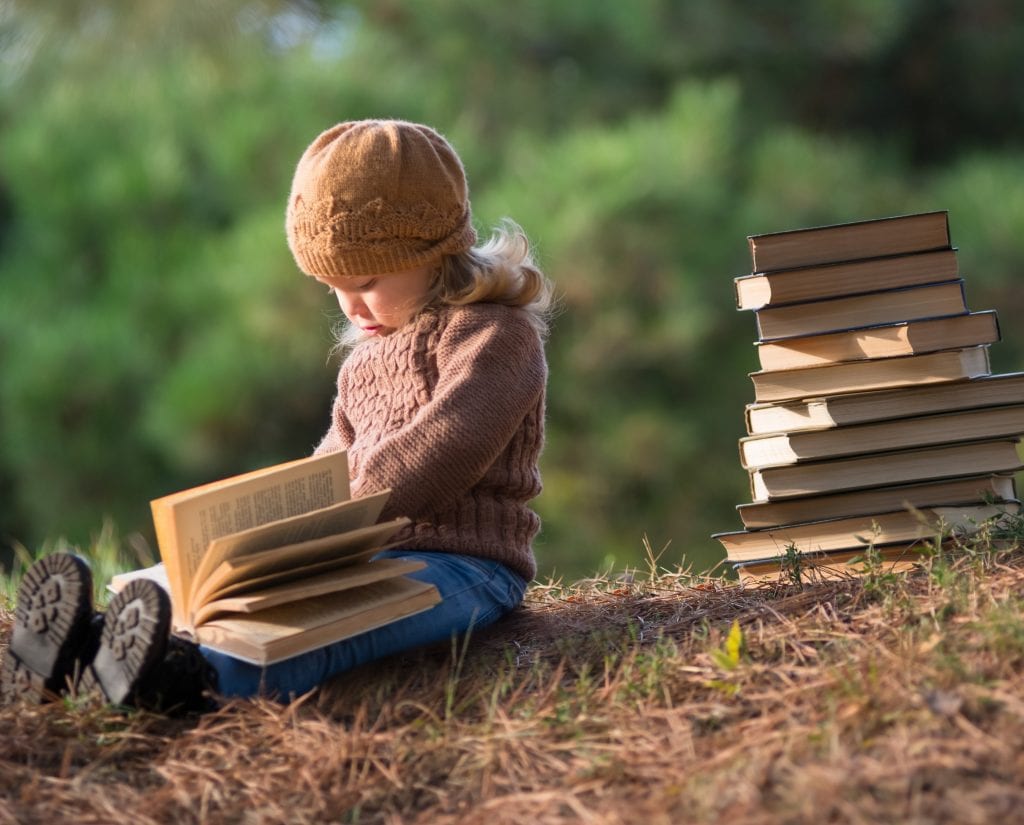 co-parenting sessions: A little girl in a brown sweater and hat is sitting on the ground and reading a book. behind her is a large stack of books