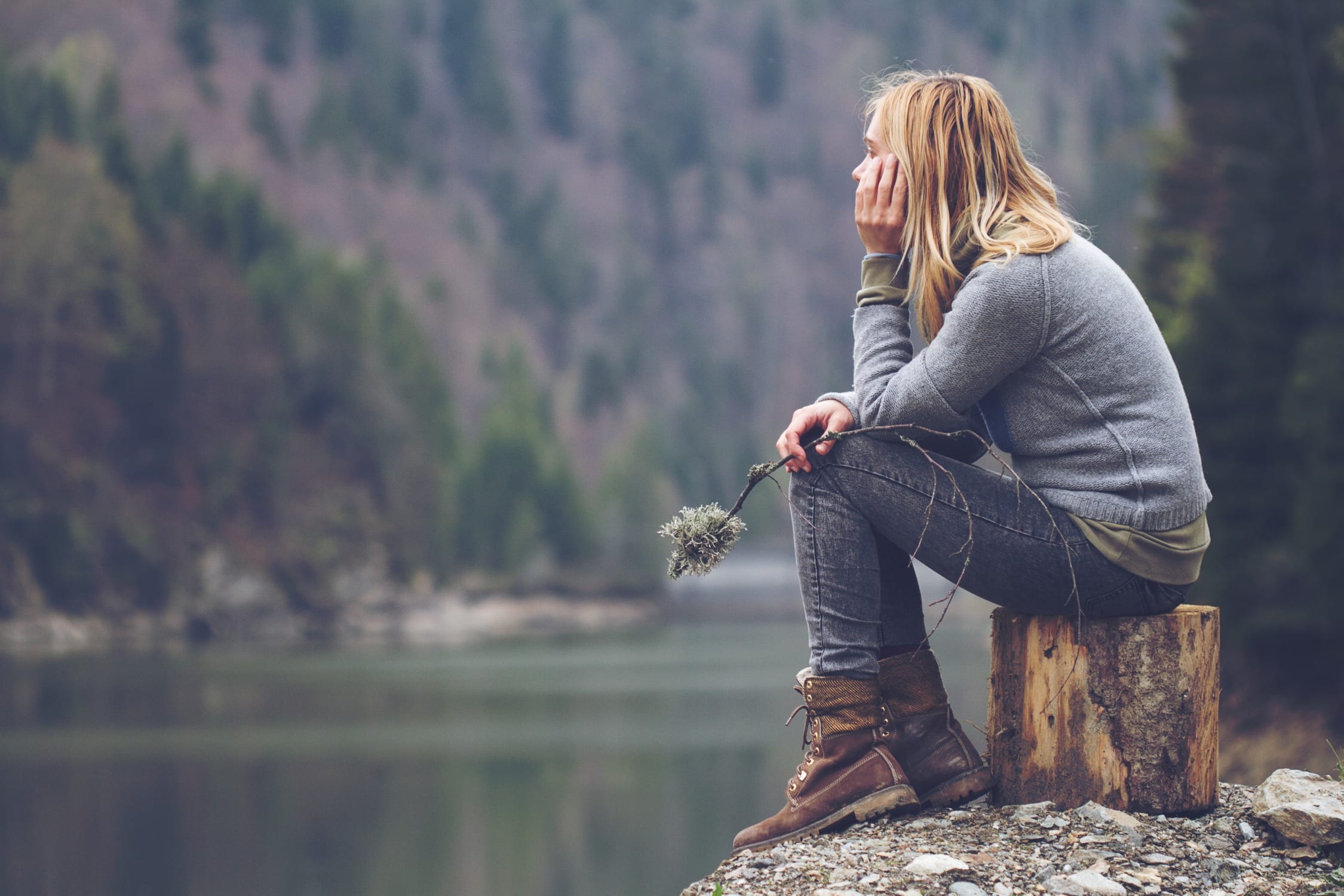 transpersonal therapy: Image of a woman sitting on a log, holding a plant and looking out over a still mountain lake.